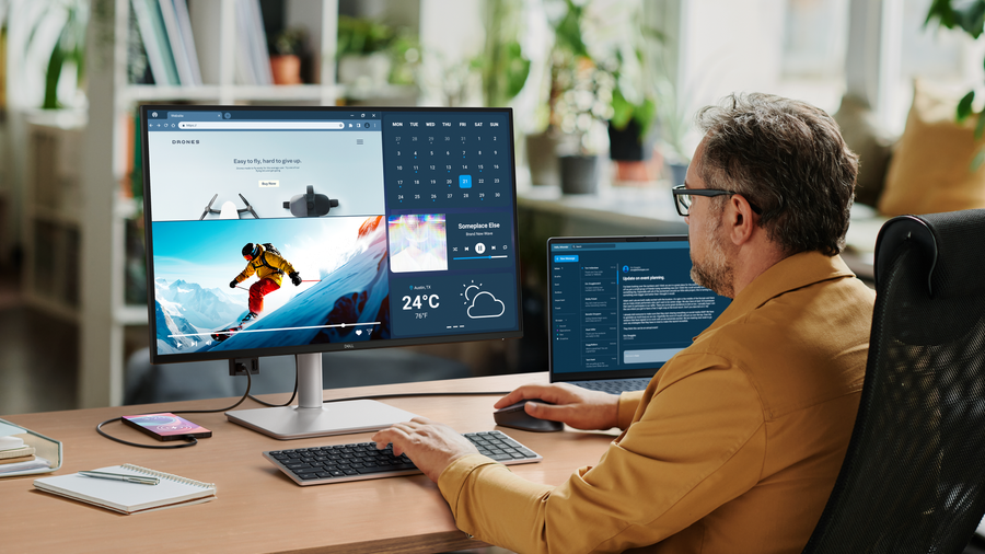 Dell S2725DC monitor on a wooden home-office desk beside a laptop, with a person using the keyboard and mouse.