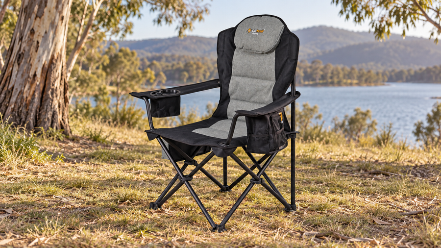 Black and grey OZtrail Big Boy Arm Chair on grass at a lakeside campsite beneath eucalyptus trees.